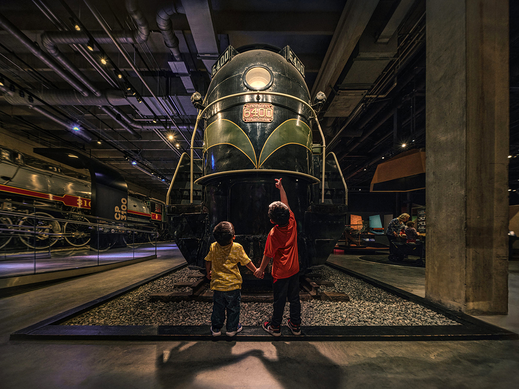 Two young children hold hands, looking up in awe at an antique locomotive.