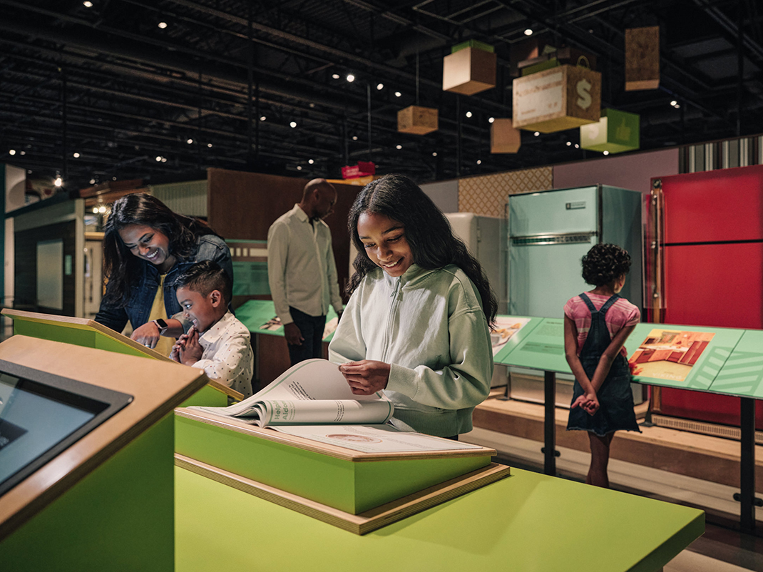 Une famille explore une exposition interactive dans un musée. Une jeune fille sourit en feuilletant un livre sur un présentoir vert. Une femme et un jeune garçon s'intéressent à une autre exposition interactive située à proximité. À l'arrière-plan, un homme plus âgé et un autre enfant observent des appareils rétro exposés, notamment des réfrigérateurs vintage colorés.