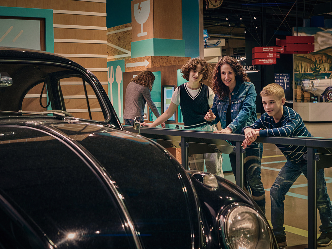 A family observing a vintage black car in a museum. An adult and two teens are leaning over a railing, looking at the vehicle with interest while another person examines an interactive display in the background.