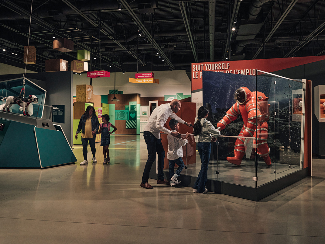 A museum exhibit featuring a family exploring displays of wearable technology. On the left, a model of a dog wearing specialized equipment is displayed. To the right, a child and an adult view a large red, futuristic diving suit within a glass case, labeled 