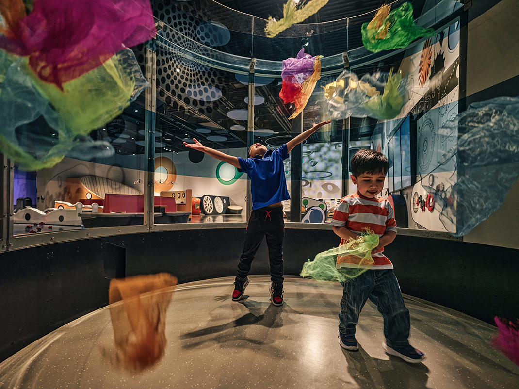 Two small children are inside of a simulated wind tunnel in a museum. One of the children is holding a green silk and the other is throwing several colourful silks into the air. In the background there are several colourful interactive stations.