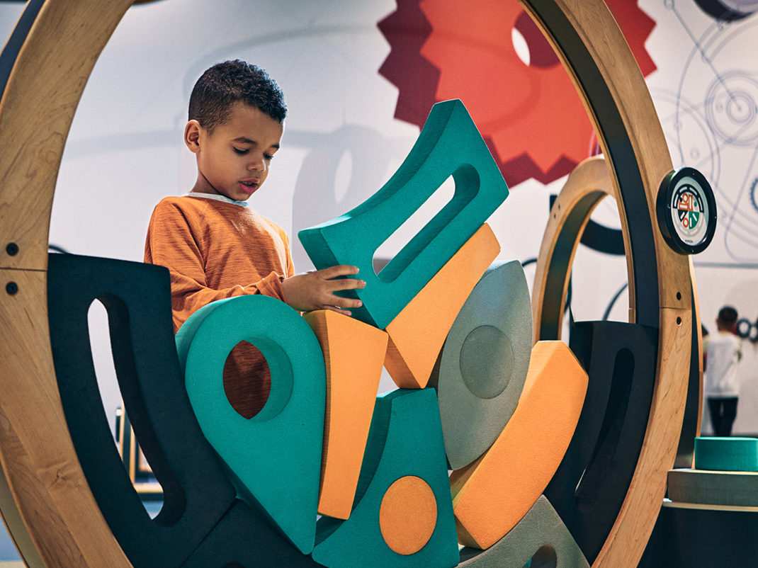 A young child wearing an orange long-sleeve shirt stands intently focused, arranging colorful foam shapes—teal, orange, and gray—within a large circular wooden frame. The interactive activity is set in a playful, hands-on exhibit space with gears and mechanical designs visible in the background