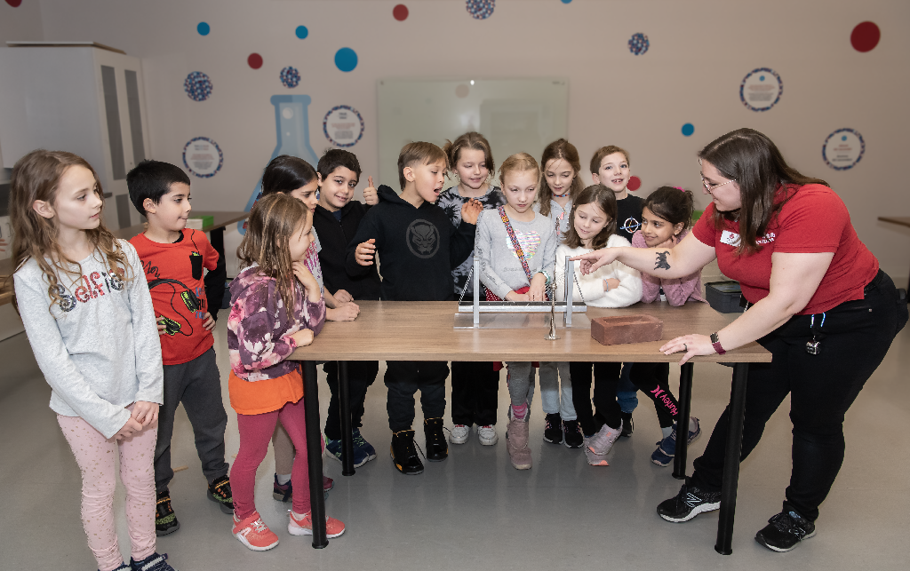 A museum staff wearing a red polo shirt is standing at a wood table as a group of children stand around it and watch her point her finger at small model of a bridge. In front of the bridge there is a brick, in the background there are colourful circles and scientific images on the walls.