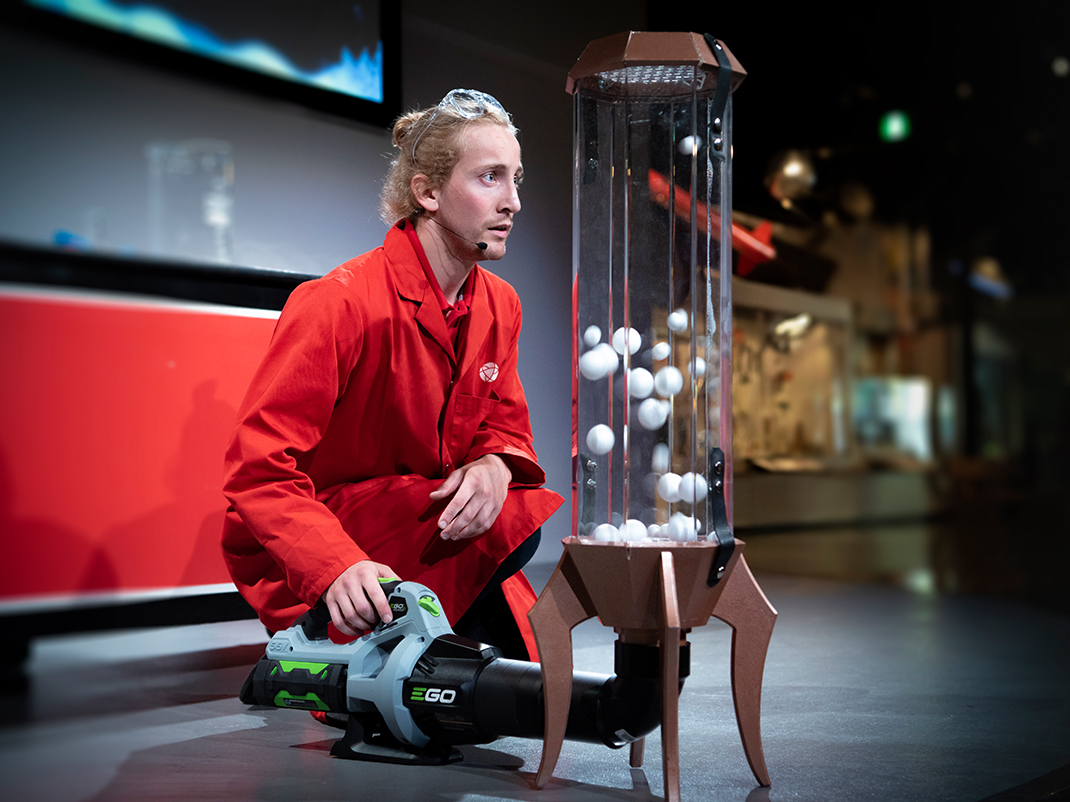 A long metal handle with a small silver ball has a spark coming out of it as it points at a Van der Graaff machine. In the top left corner there is a person in a red lab coat with protective goggles holding a stick with fire on the end of it. There are red illustrations of an explosion, a lightning bolt and a magnet on top of the image.