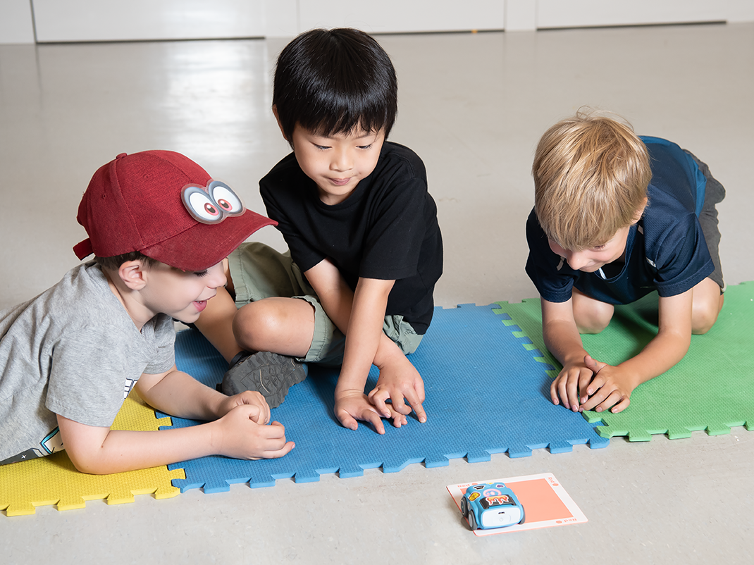 Four young children sitting on the floor watching a Sphero Indi car drive over coloured mats.