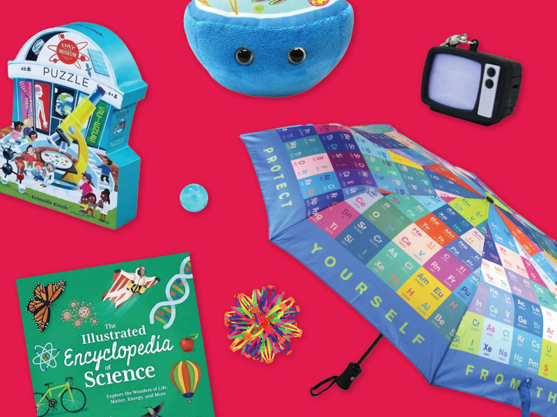 Image of various science-themed items on a bright red background. Items include a colorful periodic table umbrella with the text 'Protect Yourself from the Elements,' a puzzle box depicting a museum scene, a plush cell toy, a retro-style miniature TV, a science encyclopedia book titled 'The Illustrated Encyclopedia of Science,' a rainbow-colored stress ball, and a small blue marble.