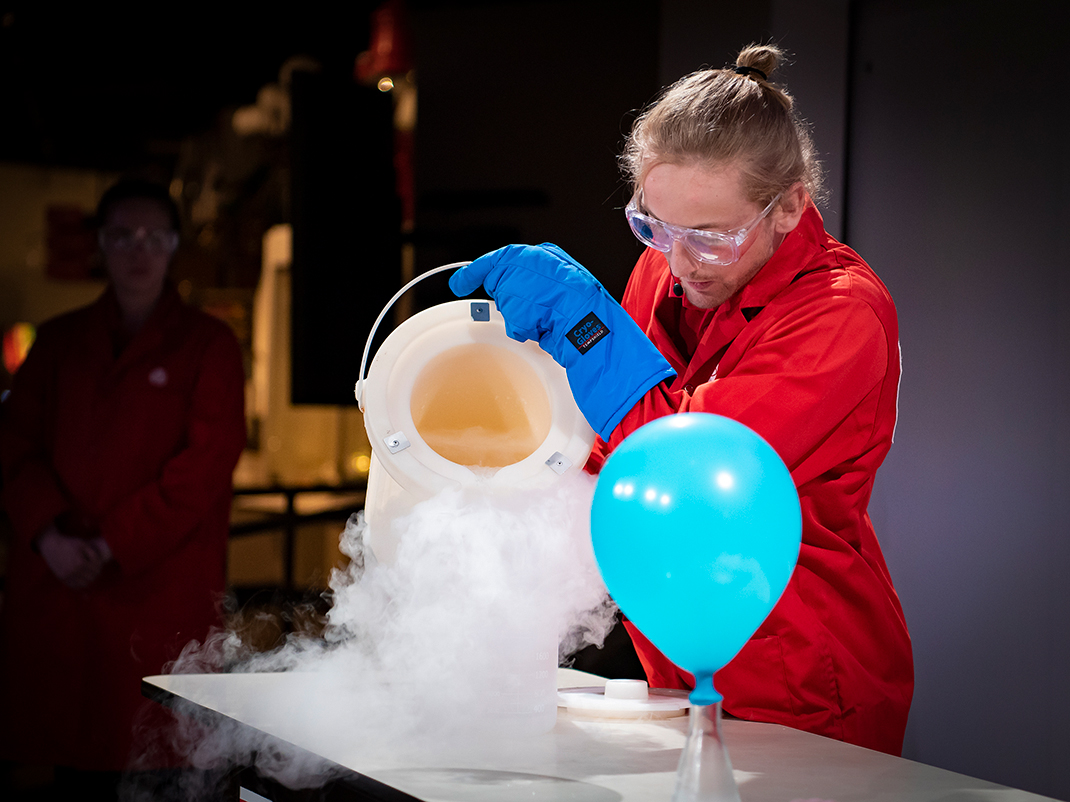 Museum guide with light skin and blonde hair in a bun is dressed in a red lab coat and pouring liquid nitrogen out of a dewar, next to it there is a blue helium balloon.