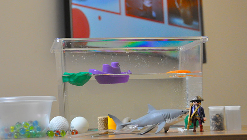 A tank of water contains a floating purple plastic boat and a green plastic rock. Outside the tank are a scattering of objects such as a plastic shark, a playmobil pirate and several golf balls.