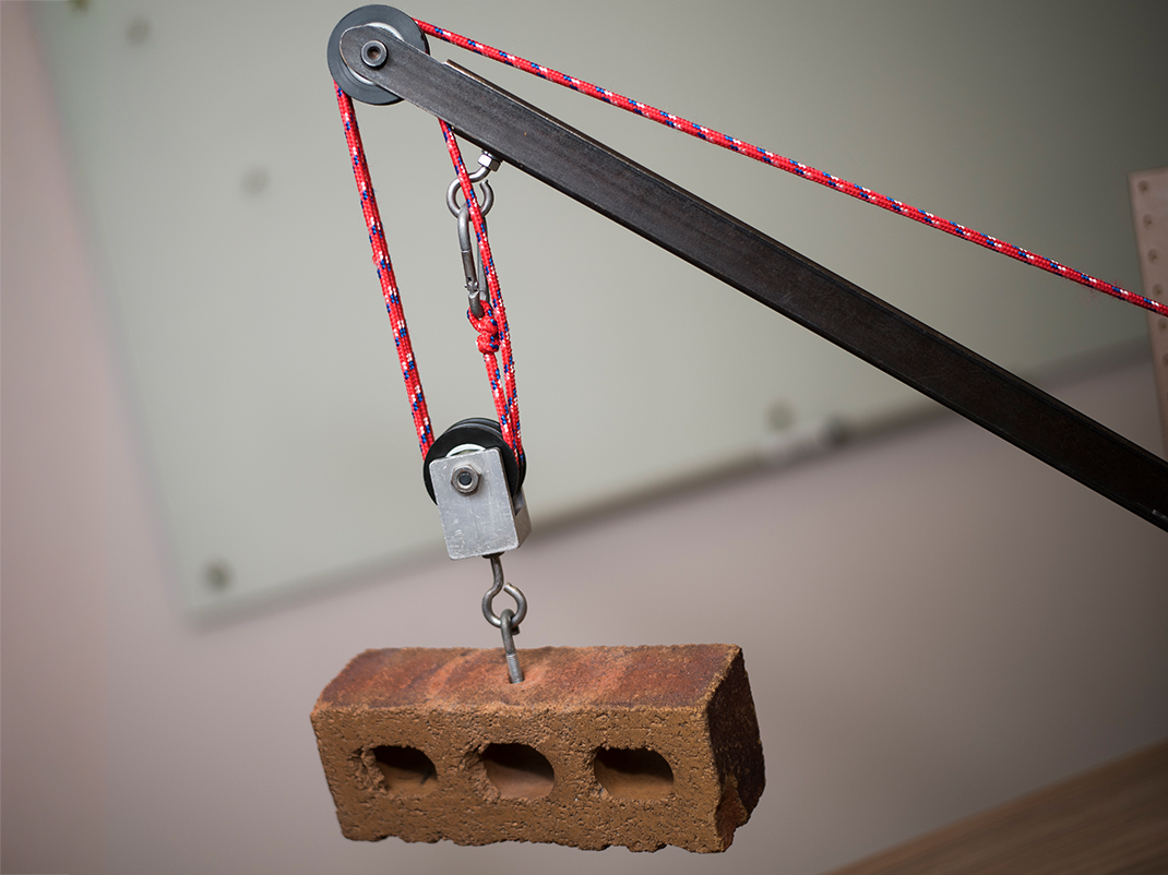 Three differently sized gears are affixed to a wooden background with holes in it. The gears are connect to a long pulley that is supporting a wooden block.