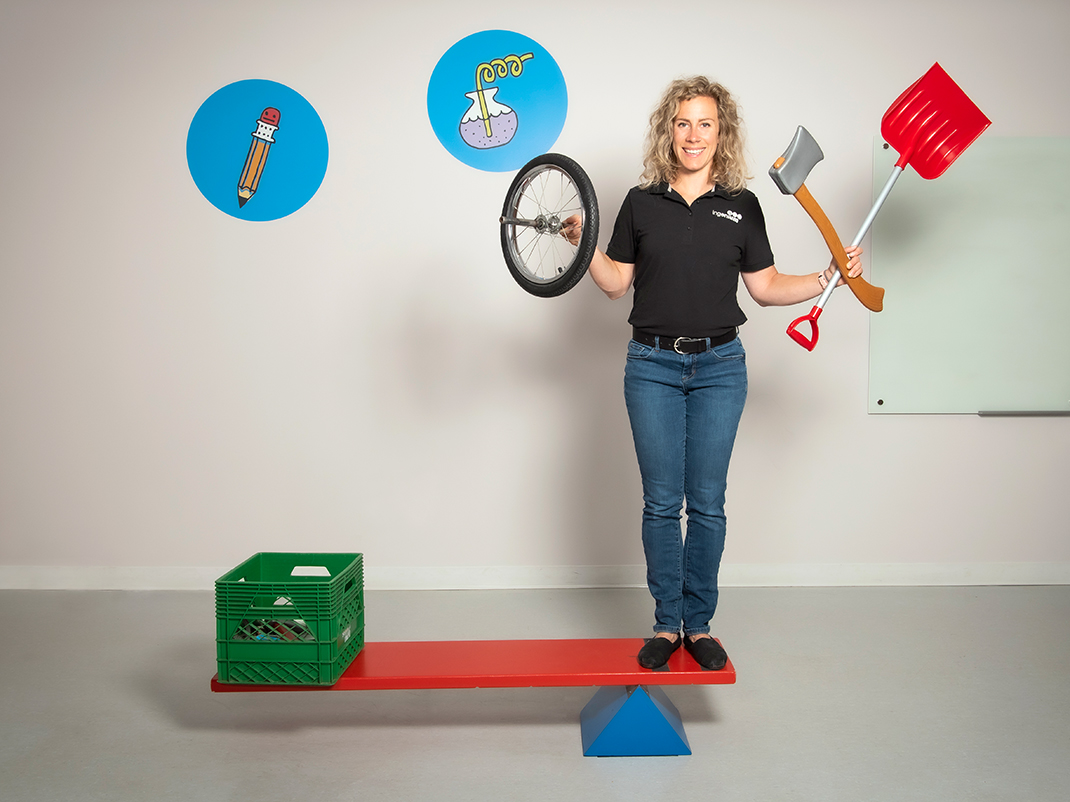 A large lever sits on the floor of a white room. On one side of the lever is a young girl, on the other side is a man. Because of the placement of each person, the lever is tilting towards the girl. A museum guide watches from behind.