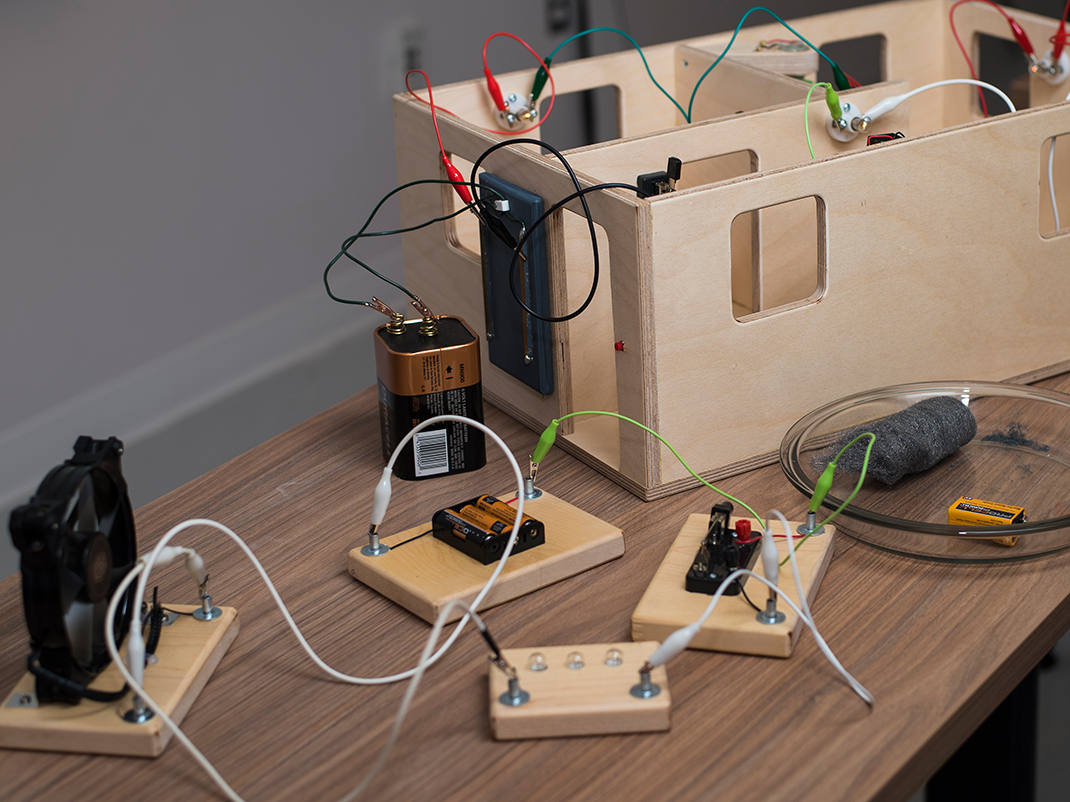 A person's hands rest on a table which is covered with rectangular boards containing batteries and wires which are attached together to form a large circuit.