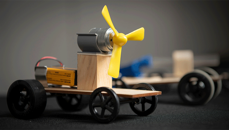 Close-up of a small wooden vehicle with black wheels and a yellow propeller affixed to a grey motor.