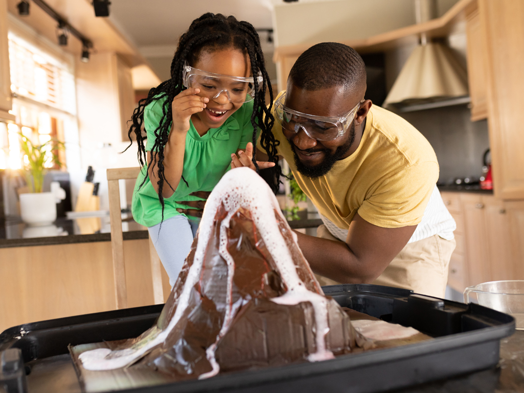 Une jeune fille noire et un adulte, portant tous deux des lunettes de protection, regardent avec enthousiasme un volcan fait maison entrer en éruption avec de la mousse dans une cuisine. La jeune fille se penche avec un grand sourire, tandis que l'adulte s'accroupit à côté d'elle, tout aussi engagé et souriant. Le modèle de volcan est posé sur un plateau, de la mousse rouge et blanche coulant le long de ses parois.