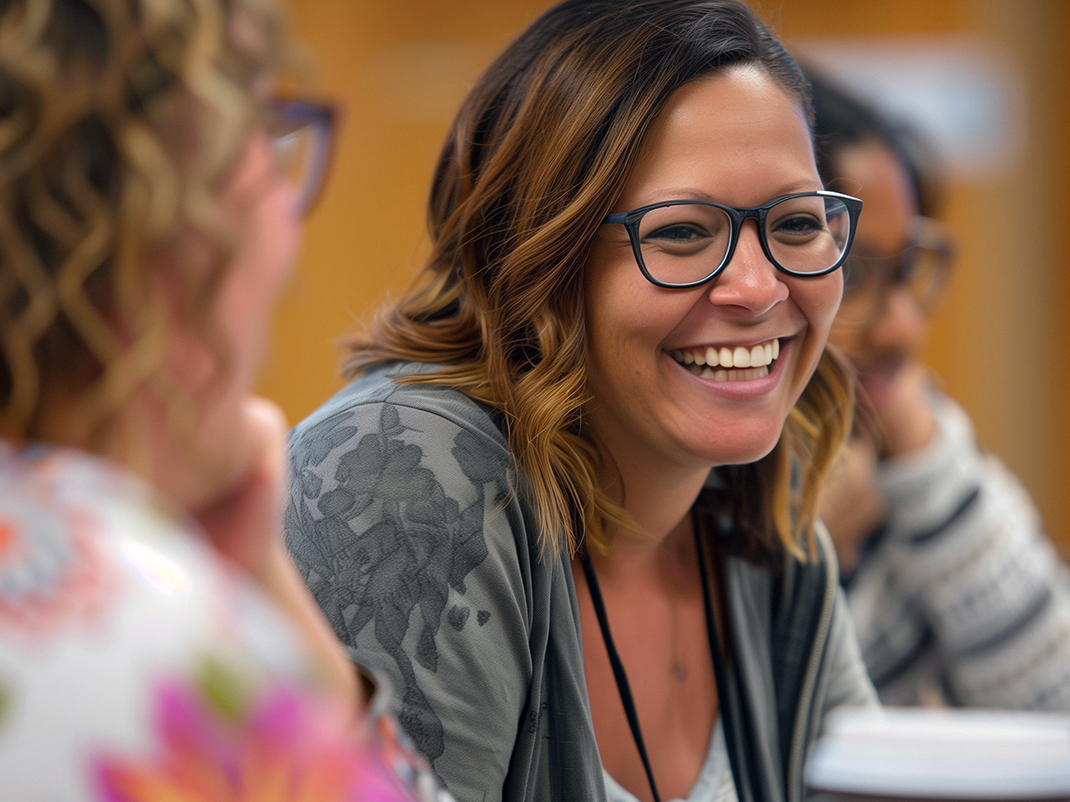 A close up of a person with shoulder length brown hair and glasses with a big smile. Blurred in the foreground and background there are two other people.