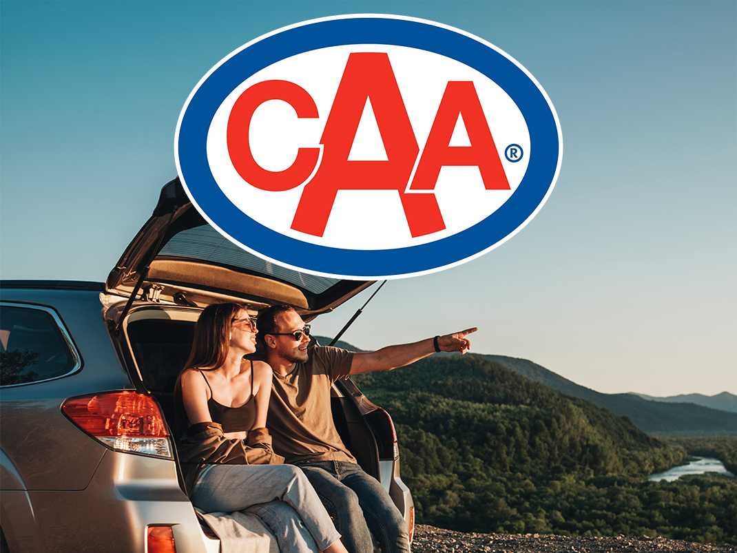 Image of a couple sitting in the open trunk of a car, parked with a scenic view of a river valley and forested hills under a clear blue sky. The man is pointing towards the horizon, while the woman smiles. Overlaid on the image is the logo of the Canadian Automobile Association (CAA), prominently displayed in the top center.