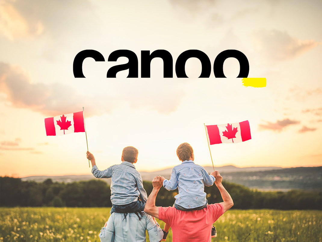 Two small children wearing blue collared shirts are holding up small Canada flags as they are sitting on their parents' shoulders. Ahead of them there is an open green field and a setting sun over small mountains.