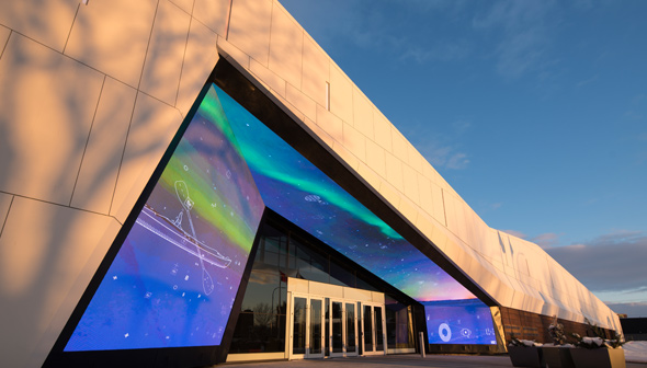 Photo de la façade du Musée des sciences et de la technologie du Canada. Le bâtiment est une forme triangulaire beige avec une série d'écrans montrant des lumières de couleurs différentes au-dessus des portes d'entrée principales.