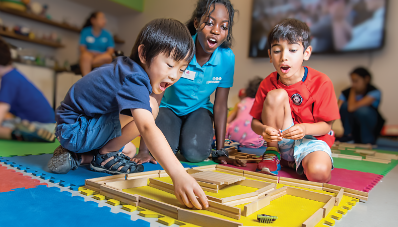 Two children and a camp counsellor wear expression of excitement as they watch a hex robot moving around a maze made of wooden blocks. In the background, other children and counselors enjoy camp activities.