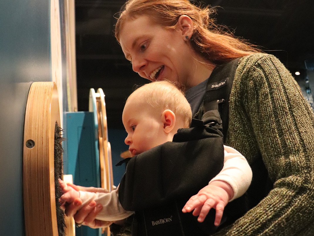 A mother with long red hair and a green sweater is carrying a baby in a forward-facing baby carrier, the mom is holding the babies hand up to a fury piece of material in a circular wooden frame.