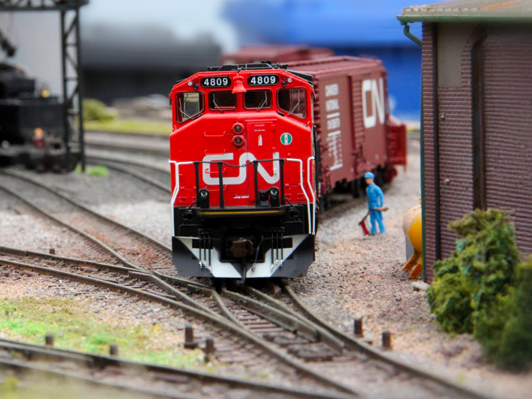 A detailed model railway scene featuring a red Canadian National (CN) locomotive numbered 4809 on train tracks. The locomotive is pulling a boxcar, and a miniature figure wearing blue overalls stands nearby. The scene is surrounded by model buildings and greenery.