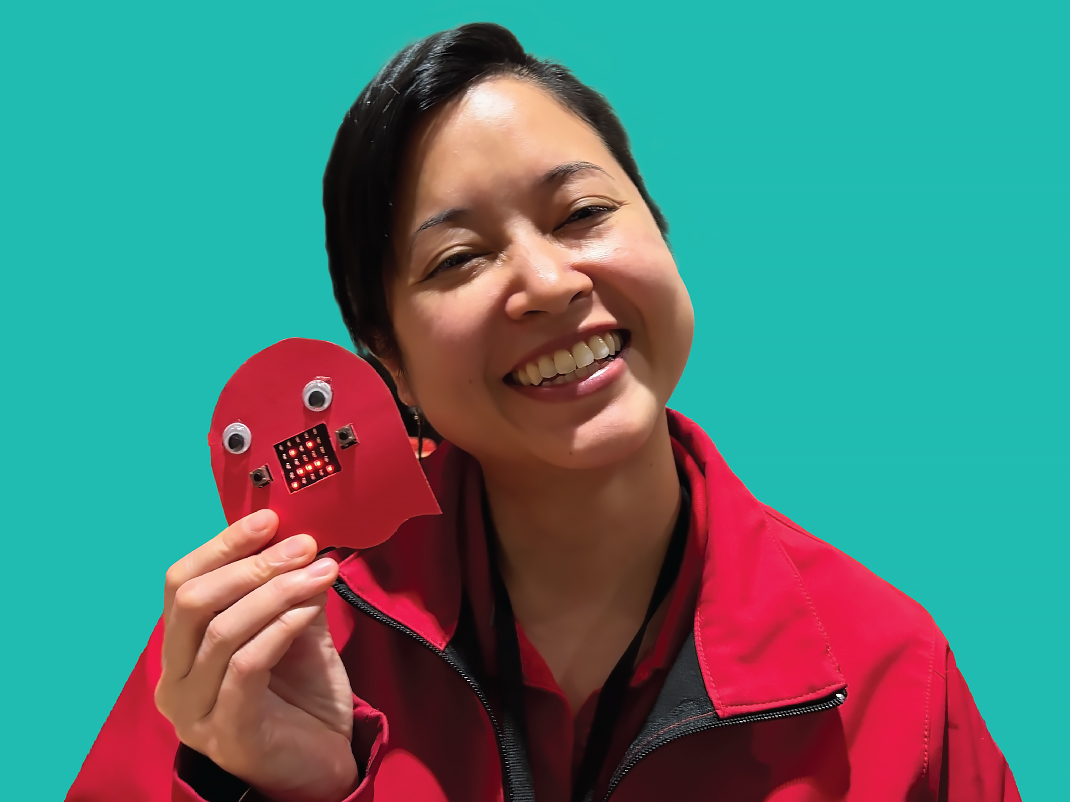 A person in a red coat holding up a red Micro:bit animal while smiling. It is set against a turquoise background.