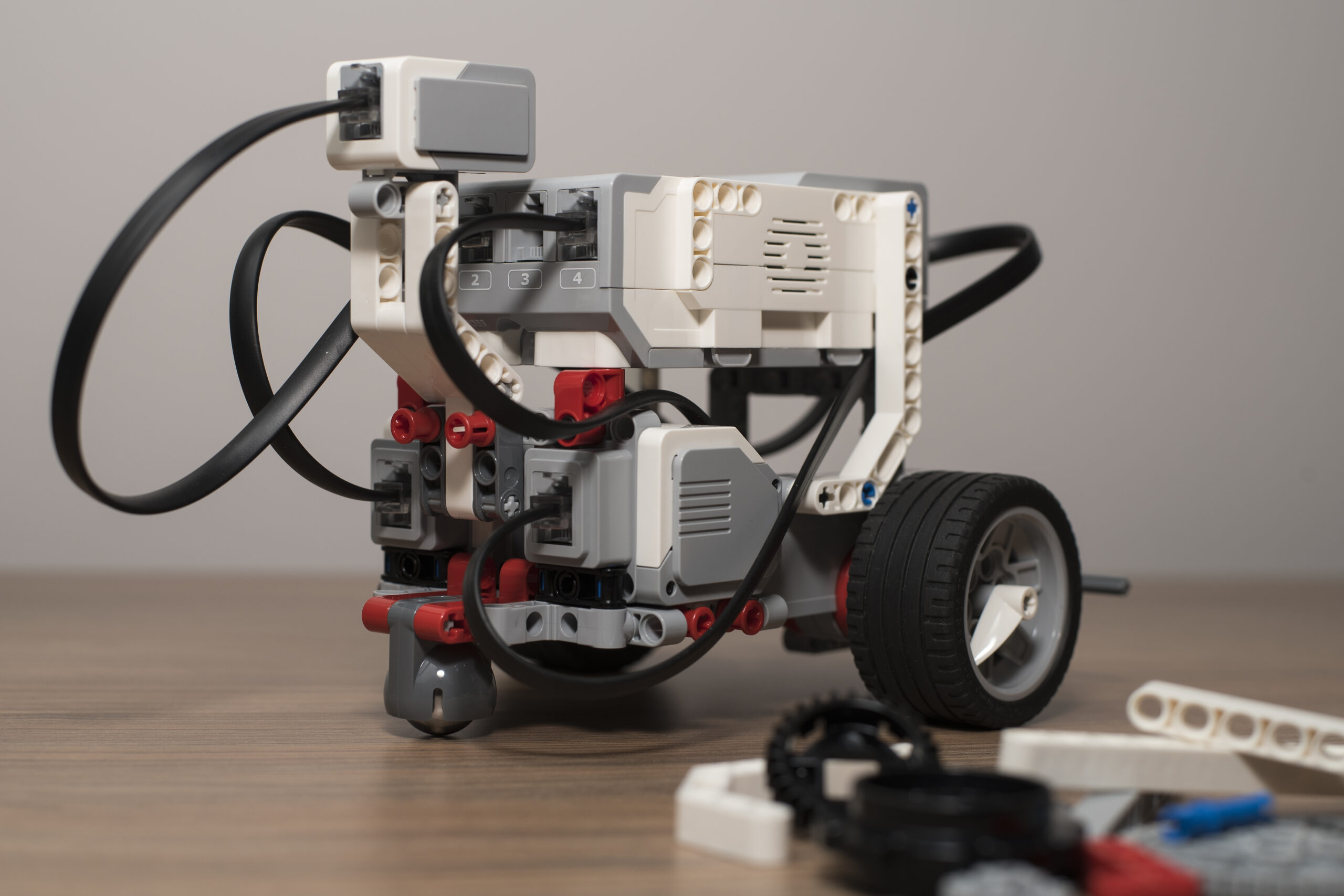 A LEGO Mindstorms robot built with white, gray, and red LEGO Technic pieces, featuring wheels, sensors, and black connecting cables. The robot is placed on a wooden surface with additional LEGO parts scattered in the foreground.