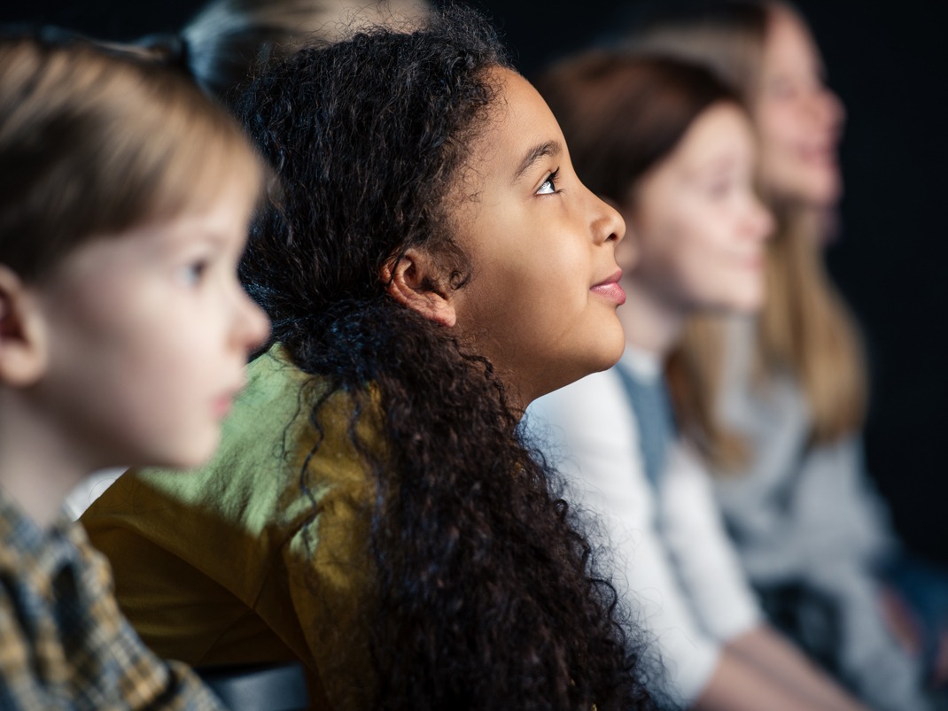 Vue latérale d'un groupe de personnes assises en rang, regardant quelque chose hors champ dans un décor faiblement éclairé.