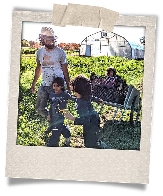 Image of a person walking in a grassy field with two smaller children. In the background we see a greenhouse.