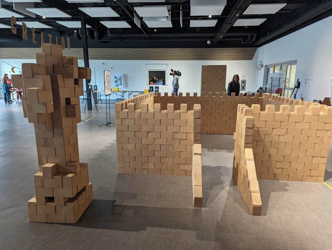 A life-size castle structure made of stacked cardboard bricks stands in an indoor event space. The castle has crenelated walls, an arched entrance, and a ramp leading up to the doorway. To the left of the entrance, a tall abstract statue, also made of cardboard bricks, resembles a humanoid figure. In the background, visitors explore the room.