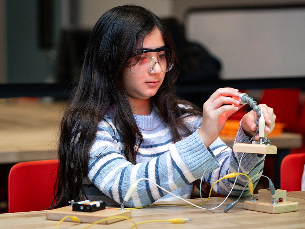 Un enfant aux longs cheveux bruns portant des lunettes de protection est assis à une table en bois sur une chaise rouge et joue avec un circuit fait maison.