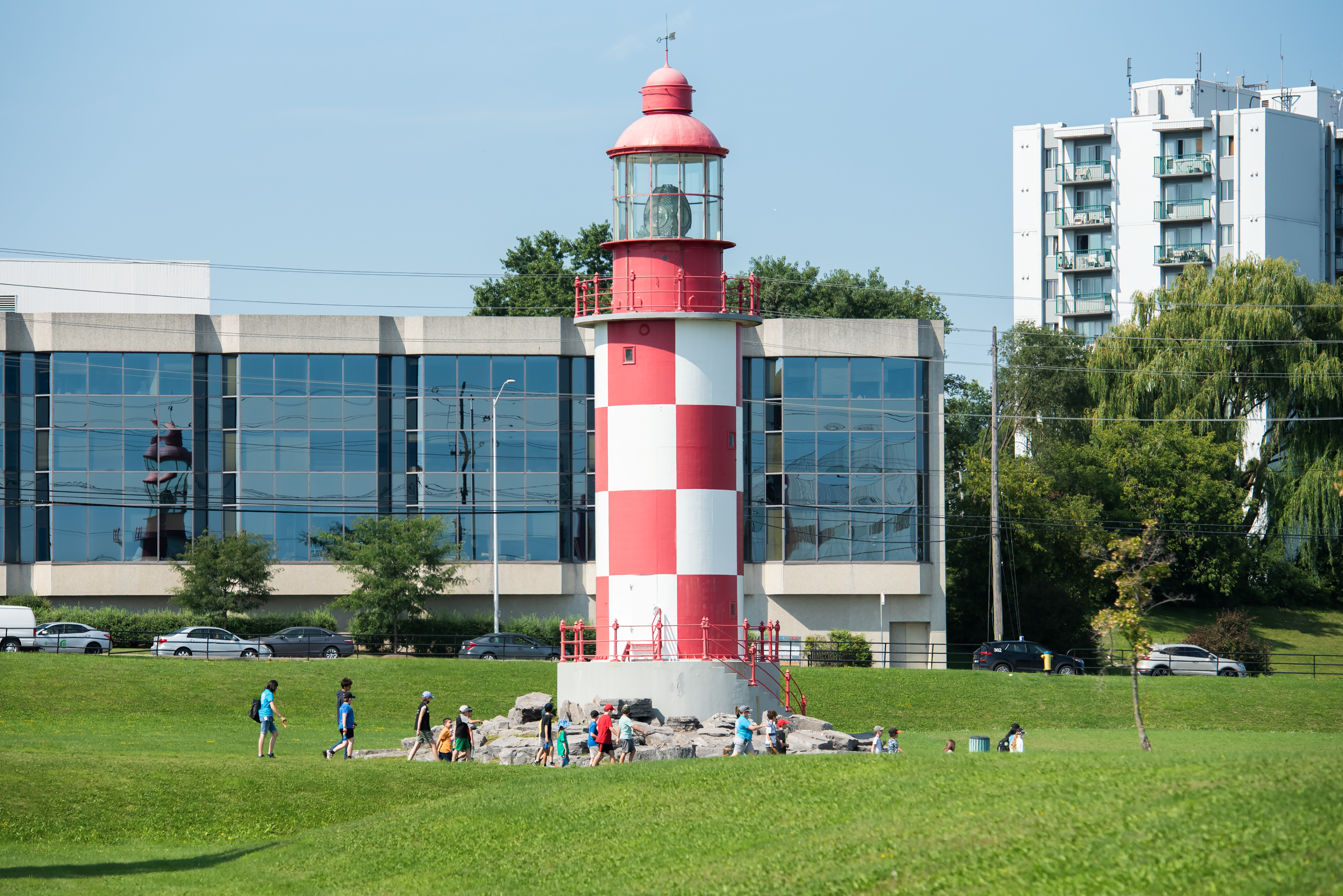 Red and white checkered lighthouse standing on a green lawn with people walking up to it. In the background there is a blue building and some trees.