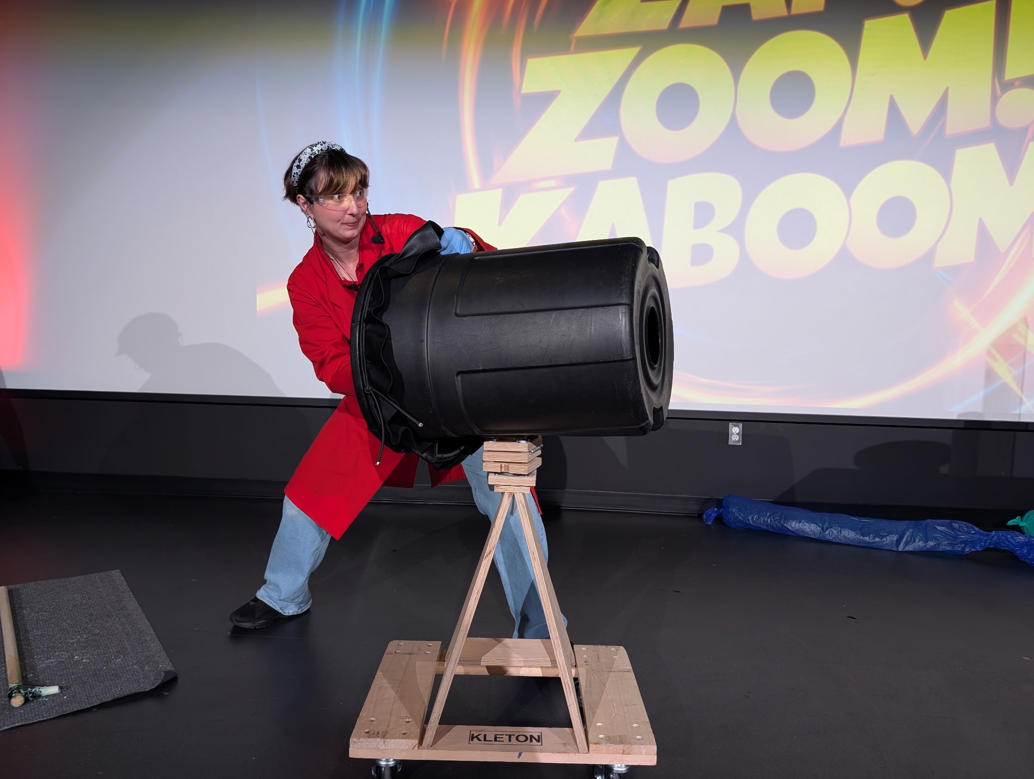 A person wearing a red lab coat and protective glasses is standing on a black stage aiming a black plastic garbage can on a wooden stand with a concentrated look on their face. In the background there is yellow block letters on a screen that say "Zoom! Kaboom!"