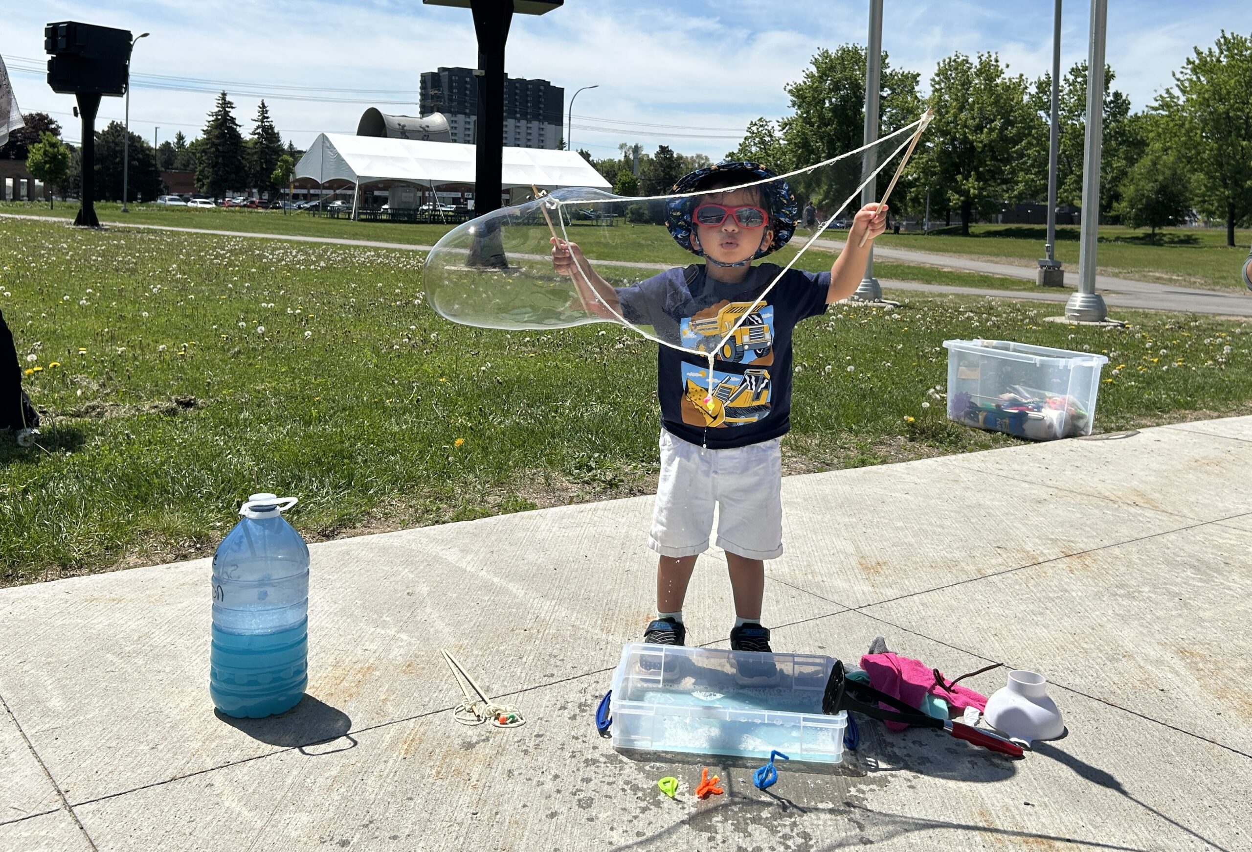 A young child is outdoors on a sunny day creating large bubbles using a rope wand. In the background there is a green lawn and a white outdoor tent.