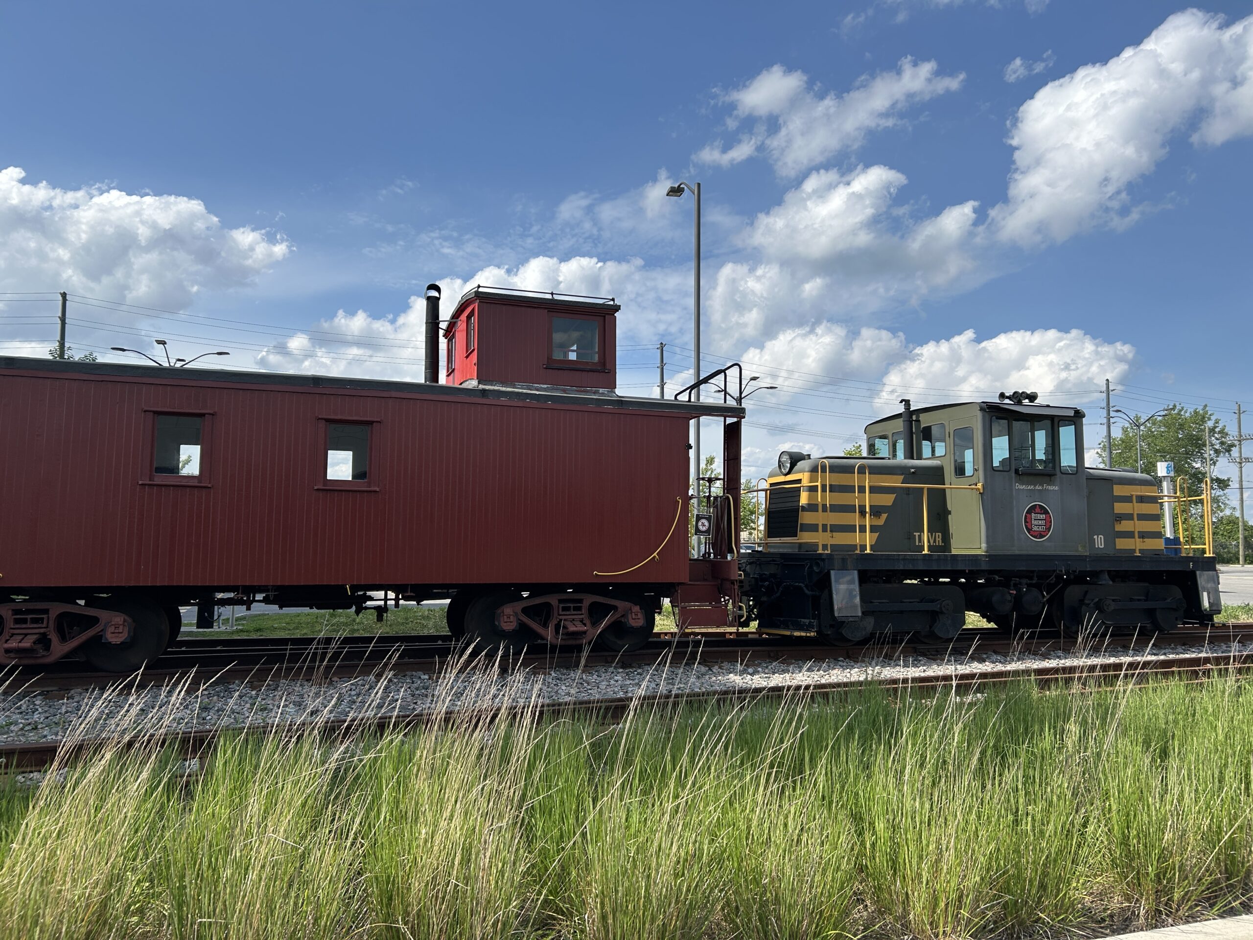 A red caboose connected to a green and yellow engine is sitting on railway track. In the forefront there is long green grass and in the background there is a blue sky with clouds.