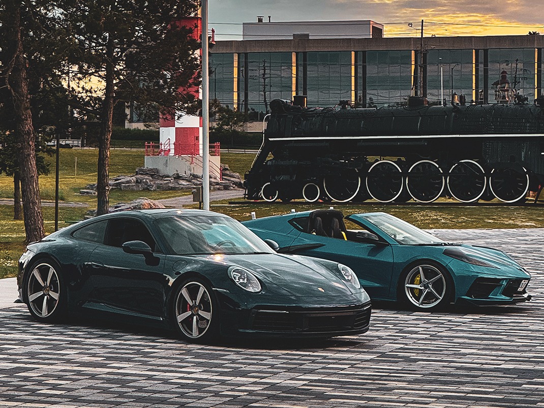 Two luxury cars, one black and one blue, are parked side by side in a parking lot. Behind them, a vintage locomotive and a lighthouse from the Museum of Science and Technology are visible.