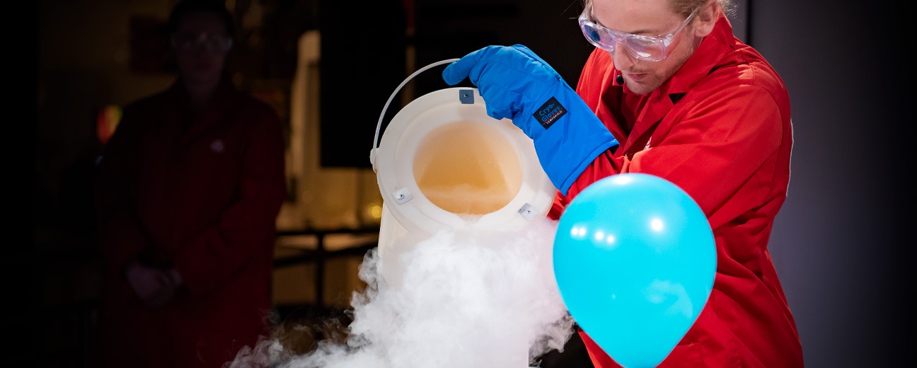 A science demonstrator wearing safety goggles, blue protective gloves, and a red lab coat pours liquid nitrogen from a container, creating a cloud of vapor next to a blue balloon.