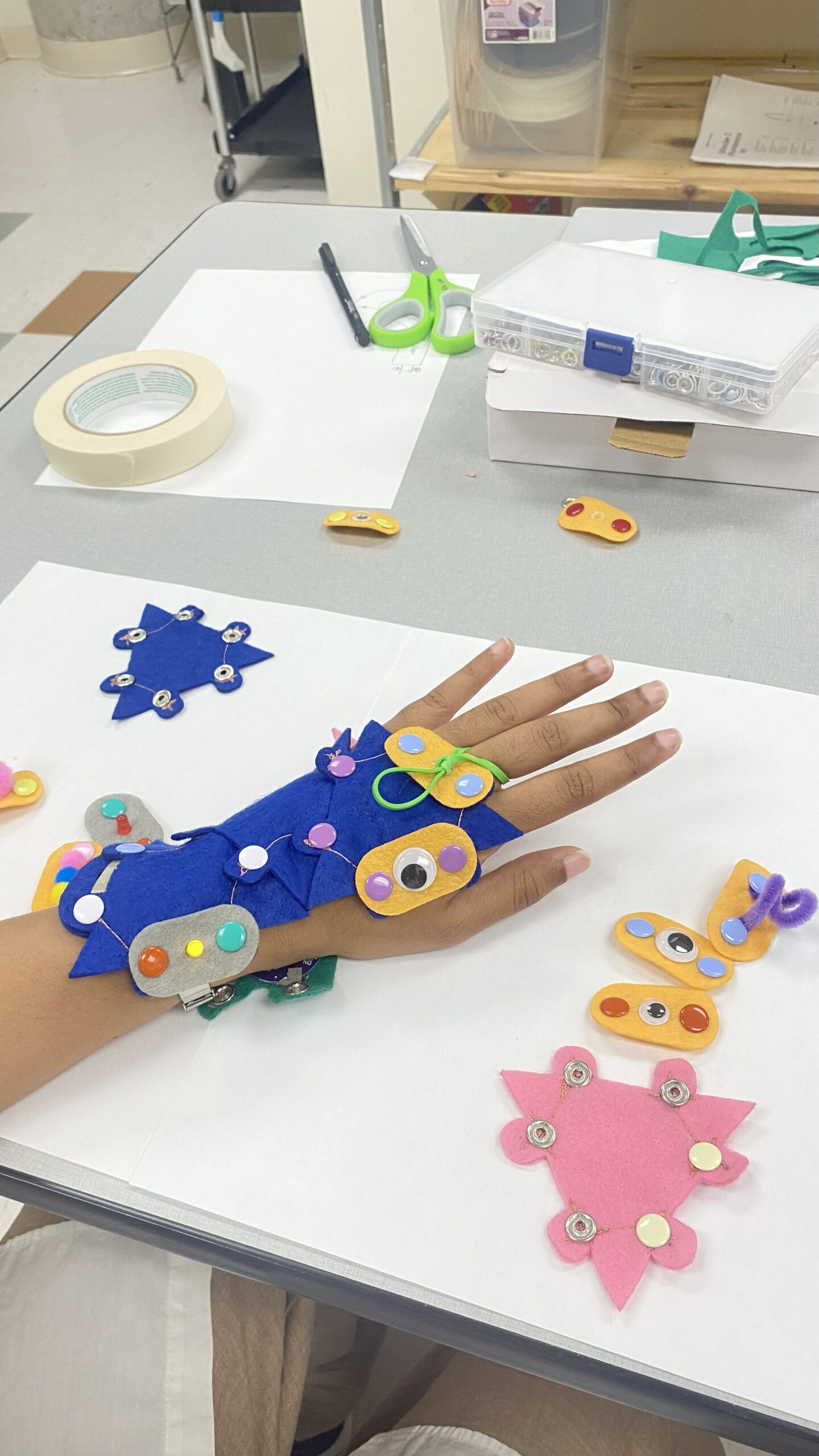 Hand wearing a blue felt wearable device decorated with colorful buttons and googly eyes, placed on a white surface with other felt pieces nearby.