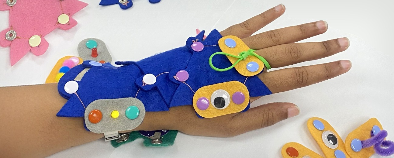 Hand wearing a blue felt wearable device decorated with colorful buttons and googly eyes, placed on a white surface with other felt pieces nearby.