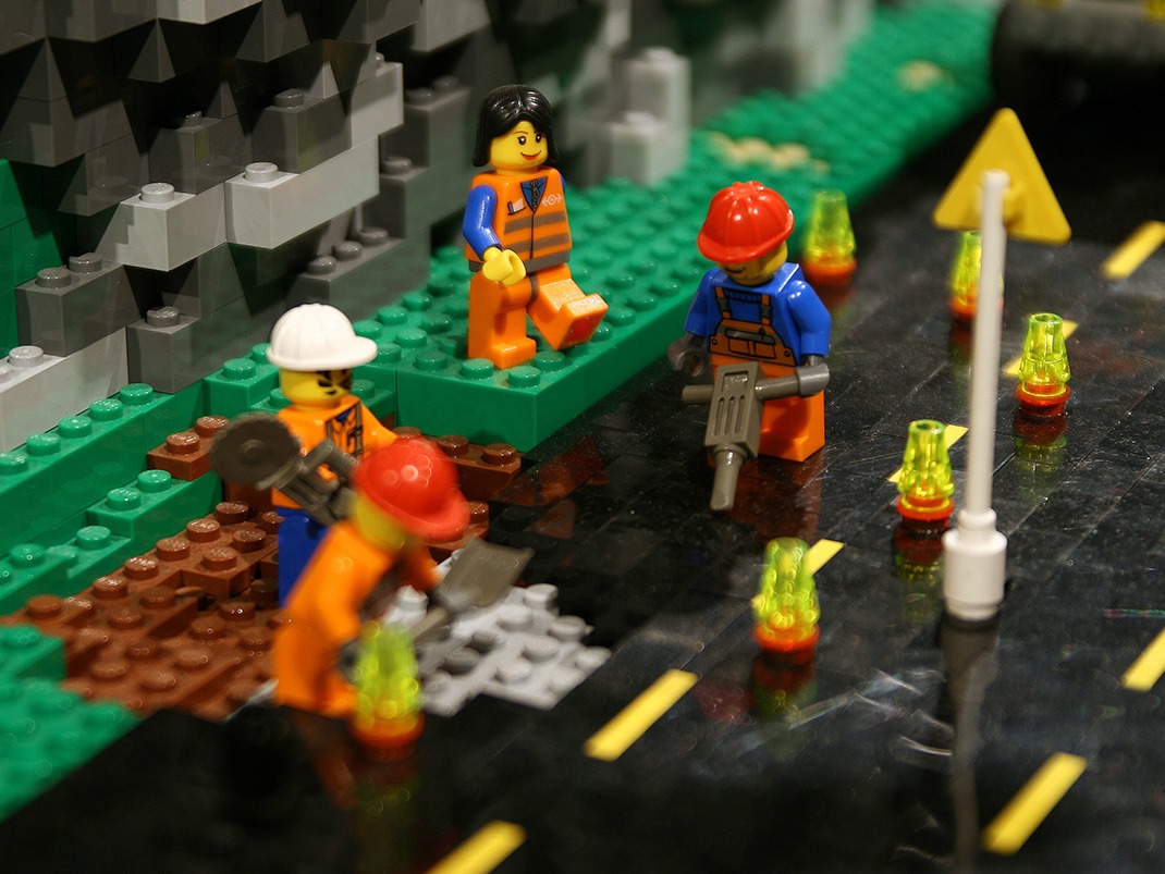 Scène de construction LEGO avec quatre personnages, portant des casques et des gilets, travaillant sur une route avec des outils. À proximité, un personnage assis et des cônes de signalisation.