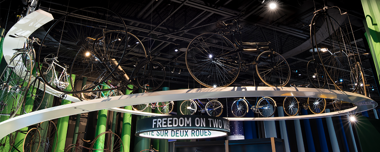 A wide shot depicts three people standing in the middle of a bike-themed exhibition. A variety of bikes are displayed on high shelving and inside a glass display case, and the words, "Freedom on Two Wheels" is visible.