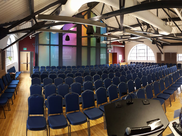 A large room with industrial-chic barn-style dark metal beams on a high and bright ceiling. The floor and walls are wood-toned and the back of the room has multi-coloured floor to ceiling glass panes and semi-circle windows. Dozens of blue single chairs are lined up facing the front of the room where there is a projector and a podium.