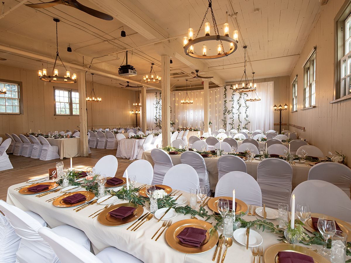 A banquet with rectangular tables of 12 people. The chairs are white, and the table is beautifully set with golden plates, cutlery and floral centrepieces.