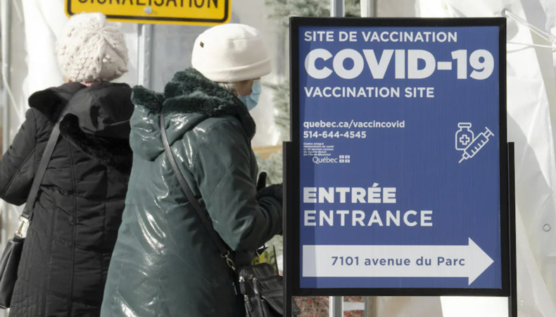 Two women in long winter jackets with white toques stand next to a COVID-19 vaccination site sign.