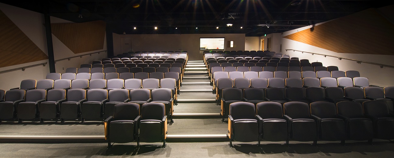 The Canada Science and Technology Museum auditorium from the perspective of the speaker at the front of the room. Rows of plush dark purple theatre-style chairs are lined up in rows going up the stairs. There are a few places at the front before the start of the stairs without chairs for accessible seating. The control room is visible at the back of the room at the top of the stairs.