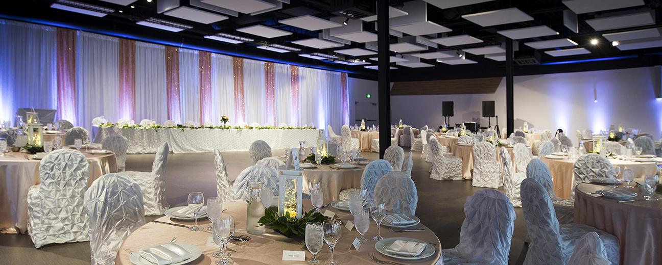 A large room with white and pink curtains and multiple round tables with floor-length pink table cloths and chairs with lush white cloth draping. The tables are set up formally for a dinner and the room is illuminated in white and blue lights.