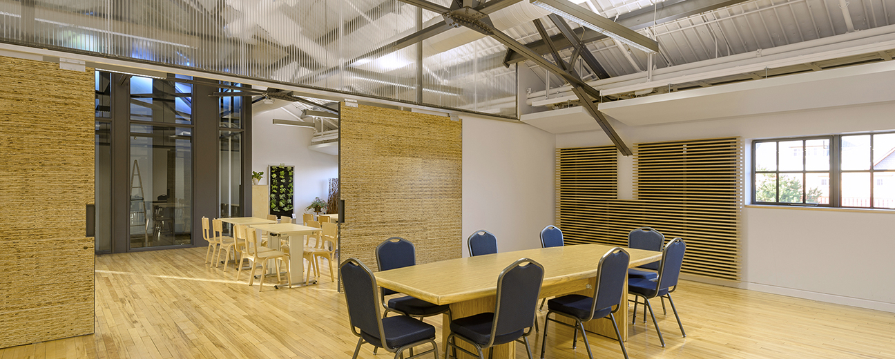 A spacious room with high ceilings and a polished wooden floor, divided by a sliding door. The front room features a table surrounded by eight blue chairs, while the back room contains two smaller tables with wooden chairs.