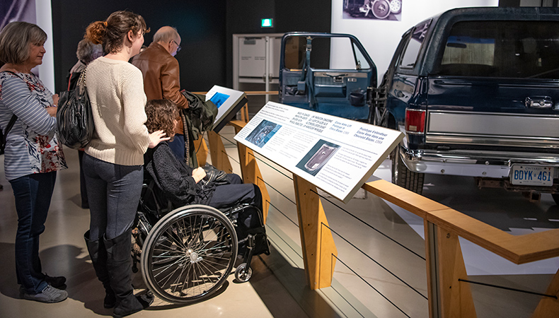 Elaine (wheelchair user) in front of an interpretation panel at the exhibit ''Moving Stories''. The artifact in the den is the first adapted vehicle Elaine ever owned.