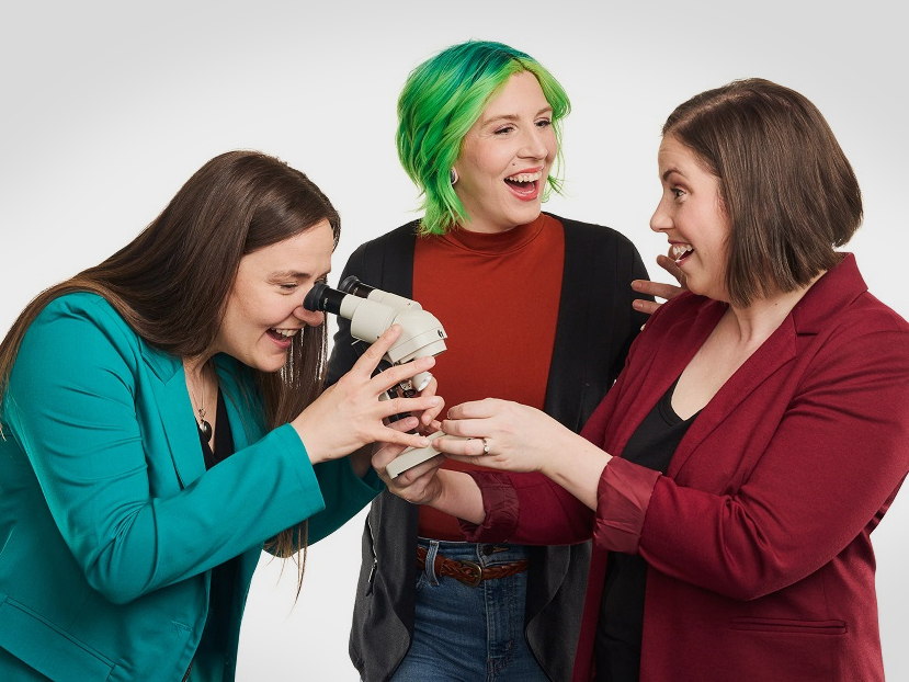 Three adults smile at one another. One peers through a microscope.