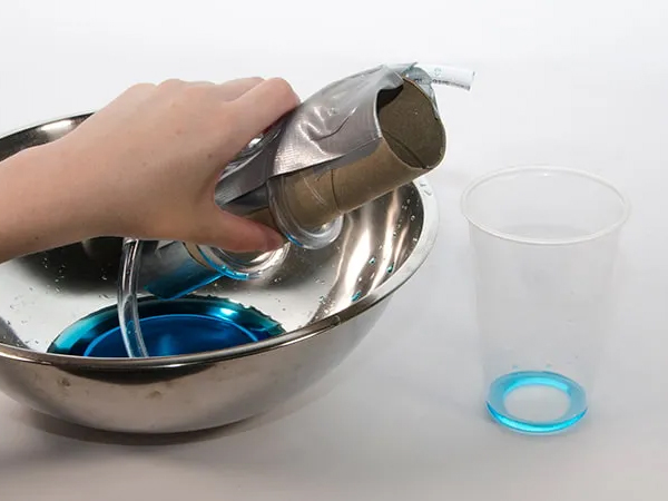 A hand reaches over a  metal bowl with blue liquid, a cardboard tube, and a plastic glass.