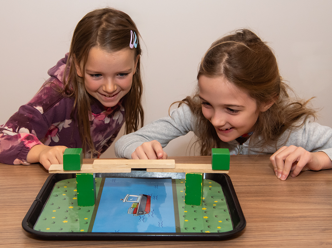 Two young girls enthusiastically playing with a balance beam activity on a table, trying to keep it balanced.